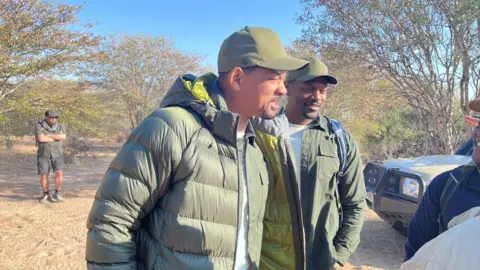 Shad Ellis Will Smith is standing with a man who looks like him and is wearing the same clothes (a green jacket and cap) in bright sunshine