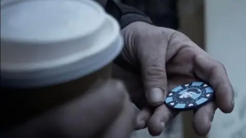 Billy Chip Close up of two hands, one is holding a take away coffee cup, the other holds a Billy Chip - a round blue and black token in the shape of a large coin, which holds a picture of a young man in a cap, surrounded by the words 'Billy Chip' and little white hearts. The hands belong to a man who is most likely homeless.