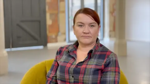 Lindsay Phelps, who is in her early 40s, is sitting in a mustard velvet arm chair in a bare, industrial-style room that is slightly out of focus behind her. She has a serious expression and dyed red hair, tied back and swept slightly across her forehead. She is wearing a navy, red and green checked shirt. 
