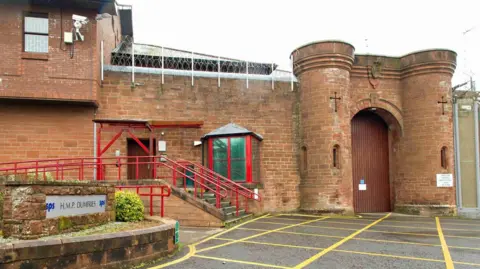 Scottish Prison Service An image showing HMP Dumfries. The building is sandstone, there is a sign to the left stating HMP Dumfries, and red railings on the stairs leading up to a brown door.