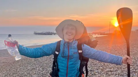 A man wearing a blue coat and holding a destroyed plastic water bottle in one hand and a paddle board oar in another. He is stood on a pebble beach at sunset with a pier in the background.