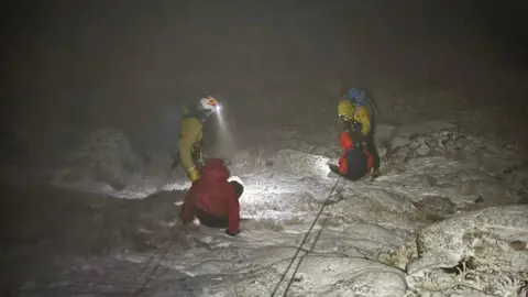 A dark photograph of a mountain rescue. Two people sitting on the snowy ground, wearing red jackets. They are attached to separate ropes and being helped by two people wearing yellow jackets, helmets and head torches. 