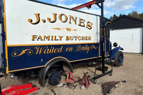 James Miller A pre-war blue van, with yellow livery around its doors, windows and edges. A sign painted on the side of the rear of the van says "J.JONES, FAMILY BUTCHER, Waiting on Daily". The rear right wheel has been removed and various tools are on the ground beside it.
