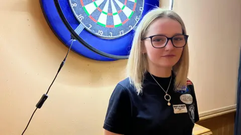 James Grant/BBC Girl wearing blue darts top poses with arms folded in front of darts board