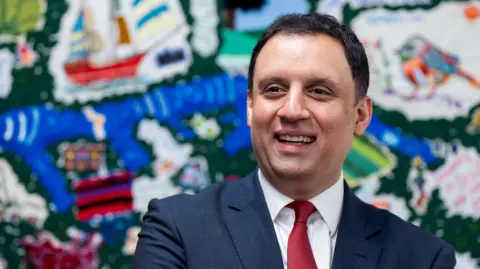 PA Media Anas Sarwar in blue suit with white shirt and red tie, stands in front of a colourful backdrop. He is smiling.