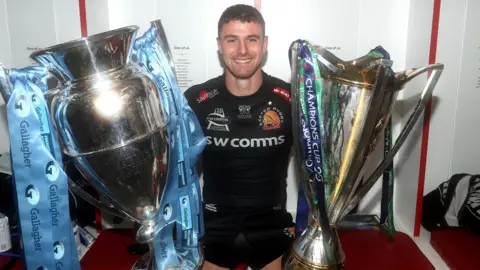 Ollie Devoto sat in a changing room wearing Exeter Chiefs kit in 2020 with the Premiership trophy on one side and Champions Cup trophy the other