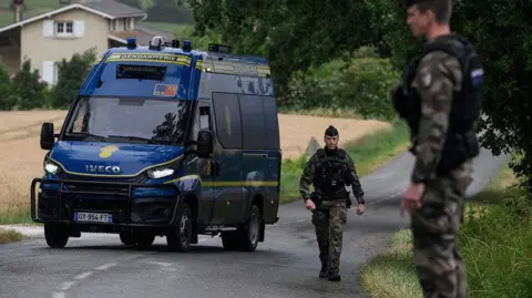 French police officers stand near their van on a road outside a french country village during the search for the missing girl. The road cuts through fields and some buildings can be seen in the background.