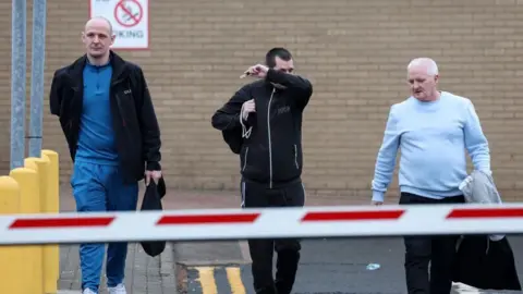 Three men leave the car park at HMP Barlinnie in casual clothing carrying black bags. The man in the middle is partially covering his face.