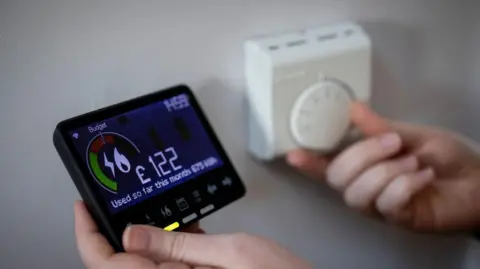 A pair of hands - one hand is holding a smart meter showing the money spent on electricity and another is adjusting a thermostat.
