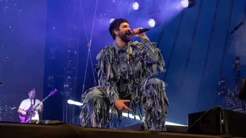 Serge Pizzorno on stage with Kasabian. He is crouched down, wearing an outfit that appears to be in tatters. A guitarist is visible in the background.