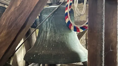 A large metal bell can be seen hanging in a wooden frame, inside a church tower. The striped bell ringing rope can be seen coiled up against it. An inscription on the bell is obscured, but the year 1886 can be made out. 