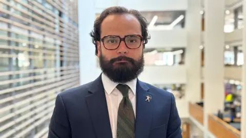 Ethan Radford is pictured wearing a navy blazer, white shirt and a green paisley tie. He has glasses and a full beard