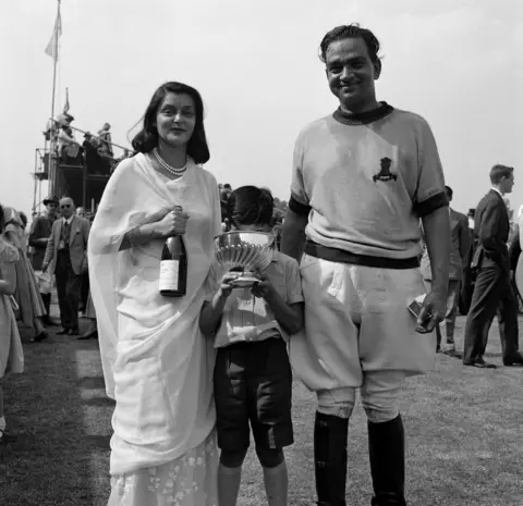 Getty Images A black and white photo of Sawai Man Singh II (1912 - 1970), the Mahraja of Jaipur, and his wife, Maharani Gayatri Devi (1919 - 2009). The Maharani is holding a bottle of champagne and their son Prince Jagat Singh (1949 - 1997) stands in between, holding a trophy at a polo event in 1957. 