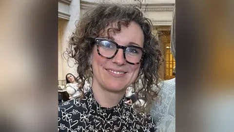 Rachel Waite with brown curly hair and brown rimmed glasses wearinng a black and white patterned blouse standing inside Liverpool's Central Library. She is smiling