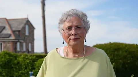 carole wyatt, a woman with short grey hair, wire frame glasses and a pale green shirt, stands in front of a hedge and a house