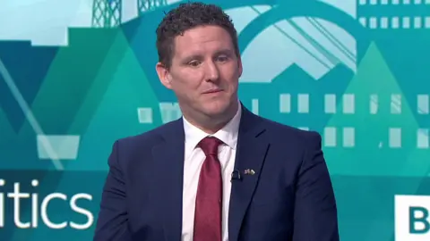 BBC Jason O'Connell, wearing a blue suit, white shirt and burgundy tie. He has short brown hair and is clean shaven. He is seen in BBC Politics Wales' TV studio during a debate on Sunday
