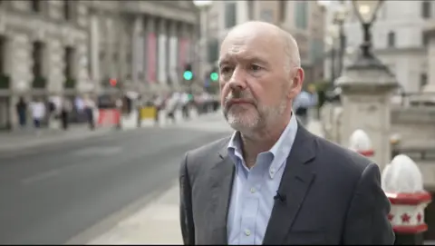 Middle aged balding man wearing a grey jacket standing in a busy street in front of historic buildings