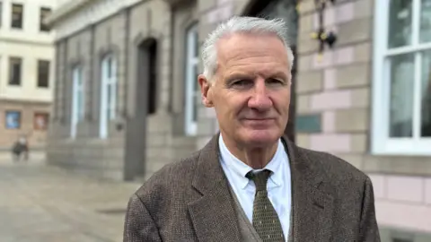 Deputy Tom Binet is standing outside a stone building smiling at the camera. He is wearing a brown and white tweed jacket over a tweed waistcoat with a black and gold patterned tie over a white shirt. He has short grey hair. 