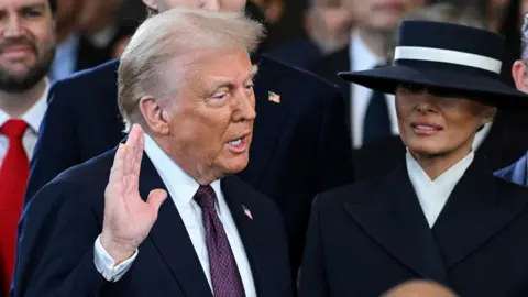 Donald Trump stands with his right hand raised and Melania at his side