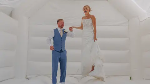 Georgia Anne Photography A woman wearing a white wedding dress stands next to a man in a sky blue suit on a white bouncy castle.
