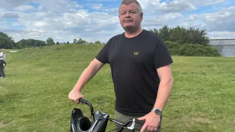 Simon Thake/BBC A grey-haired man in a black T-shirt stands on the grass with a black BMX bike