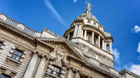 Low-angle view of a historic stone building with ornate columns and carvings, featuring a large domed roof topped by a golden statue against a bright blue sky.