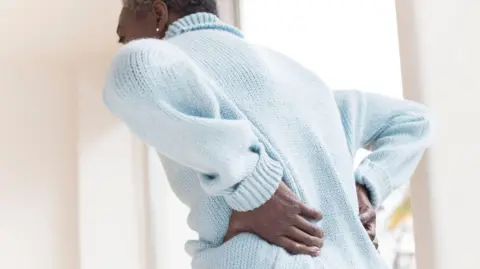 A woman wearing a pale blue jumper is seen from the back. She is leaning forward and has her hands placed on her lower back.