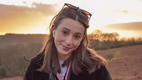 Hollinson family Darcy smiling at the camera. She has long brown hair and is standing in a field with the sun going down behind her. There is an orange glow around her. She has earpods in and sunglasses on her head.