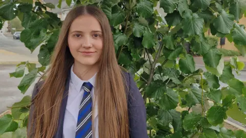 Bedford Giving Dorotea, standing by a tree, with a green bush behind her, with long straight hair, wearing a tie, shirt and blue blazer. She is smiling at the camera.
