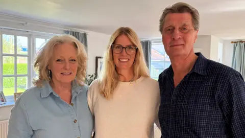 Two women and man smiling at the camera. The woman on the left has short blonde hair and is wearing a blue denim top. The woman in the middle has wavy blonde hair and has glasses. She's wearing a cream jumper. The man on the right has short brown hair and is wearing a checked shirt. Behind them are windows which have blue curtains on either side. 