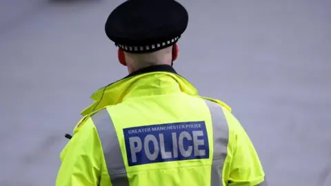 PA Media A police officer in a reflective yellow coat and wearing a black cap, with the words 'Greater Manchester Police' on the back, stands with his back to the camera