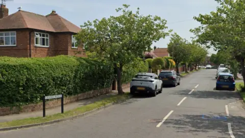 A quiet residential street with trees along the verge and cars parked up