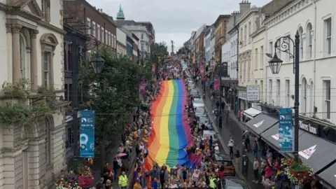 Una gran multitud, sosteniendo una bandera arcoíris, marcha por la calle Shipquay de Derry, mientras otros se alinean a ambos lados de la calle para permitir la marcha.