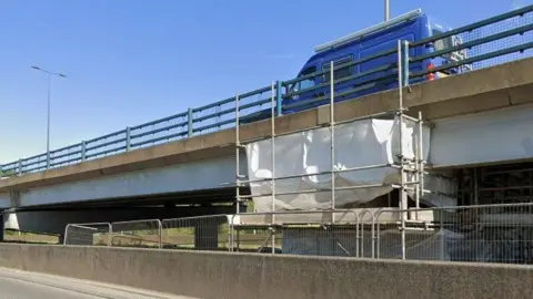 The underside of the A19 flyover has metal fencing and scaffolding in place underneath the section of road which has a blue van driving over it.