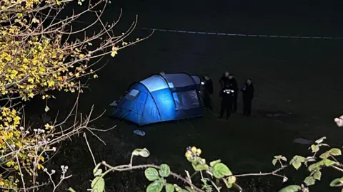 BBC/Grace McGrory Six uniformed police officers on a field next to a tent, which is lit inside