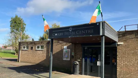 James Grant/ BBC A square brick building set next to a green bank with trees, with a concrete car park in front. The building has a black wooden awning with pillars at the front and displays a black sign saying 'Corby Irish Centre' in silver lettering. At the two front corners there are Irish flags, in orange, white and green colours. There is a blue sky in the background.