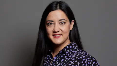 UK Parliament A portrait of Sarah Owen against a grey backdrop. She has black long straight hair, parted in the middle, and brown eyes. She is wearing a black shirt with purple shapes on it.