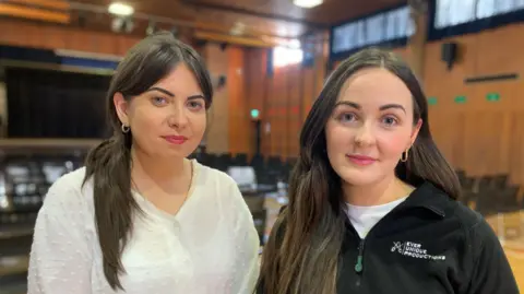 Martina McAfee and Hannah Reilly look into the camera, both have long brown hair, Martina's is tied up in a ponytail, she is wearing a white shirt and Hannah is wearing a white t-shirt with a black half-zip fleece.