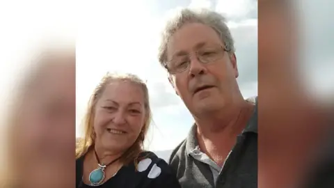 A woman with long blonde hair and her partner, a man taller than her with curly grey hair, in a selfie-style photo. The woman is smiling and is wearing a black top with white spots and a large turquoise stone necklace. The man has his mouth agape and he is wearing a dark grey polo shirt. The sea is behind them and the sun is shining.