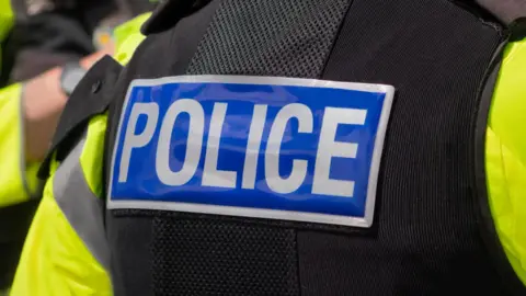 Image of the back of a police officer who is wearing a florescent top and a bullet proof vest with POLICE on the back. 