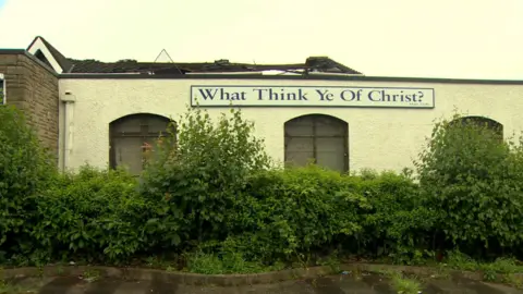 A roof of a building is damaged with sections missing. A sign on the building says, what think ye of Christ. Bushes are surrounding the building. 