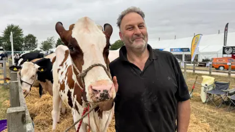Stuart Wood, who has short grey hair and facial stubble, wearing a black, short-sleeved polo shirt. He is stood in an enclosure holding a brown and white cow, smiling at the camera. Cows can be seen in the background. 