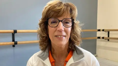 BBC Anne Sutton has shoulder length brown hair and is wearing glasses and smiling at the camera. 