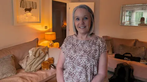 Joy Munns has short grey-blonde hair. She is wearing a cheetah print top and is standing in front of two peach coloured sofas