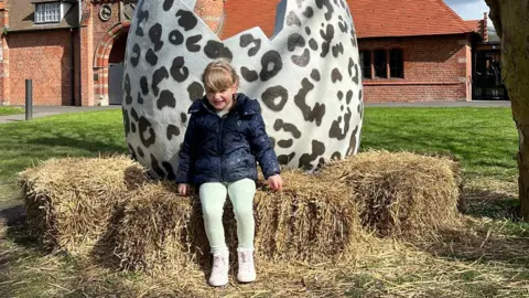 Turner Family Emilee wearing a dark blue coat, light green leggings and pink trainers sits on bales of straw with a giant black and white cracked egg sculpture behind her