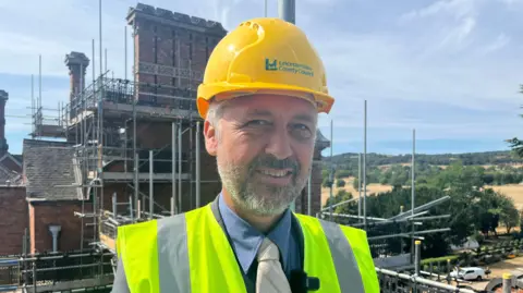 A man in a yellow hard hat and high-vis vest stands in front of scaffolding on a chimney.
