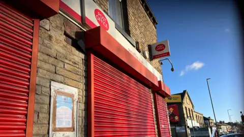 Aisha Iqbal/BBC A post office on a busy high street. The red shutters are all down and there is a closure notice visibly posted on the wall.