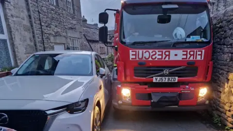 A fire engine is driving down a narrow street but has got stuck between a wall on one side and a white parked car on the other. The headlights of the fire engine are on. There are houses on the other side of the car.