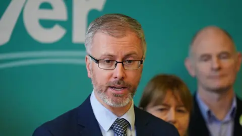 PA Media Green leader Roderic O'Gorman, with party candidates, speaking during a press conference at the Irish Architectural Archive, Dublin. He has greyish hair and brown eyes. He is wearing glasses, a dark blue blazer, light blue shirt and check tie.