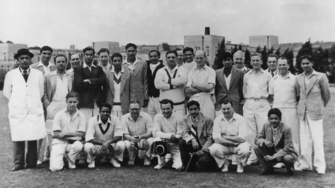 Lansdown Cricket Club A mixture of Pakistani and English cricketers in a black and white photo, wearing cricket outfits including V neck jumpers. They are on a pitch with tower block buildings in the distance.